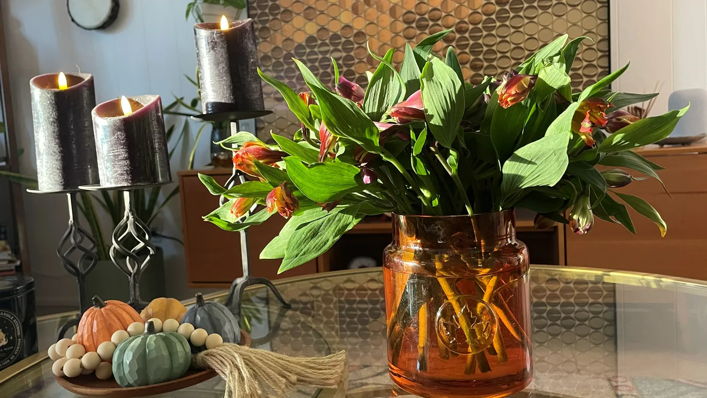 Alstroemeria in an amber jar on her table, candles lit beside little pumpkins.