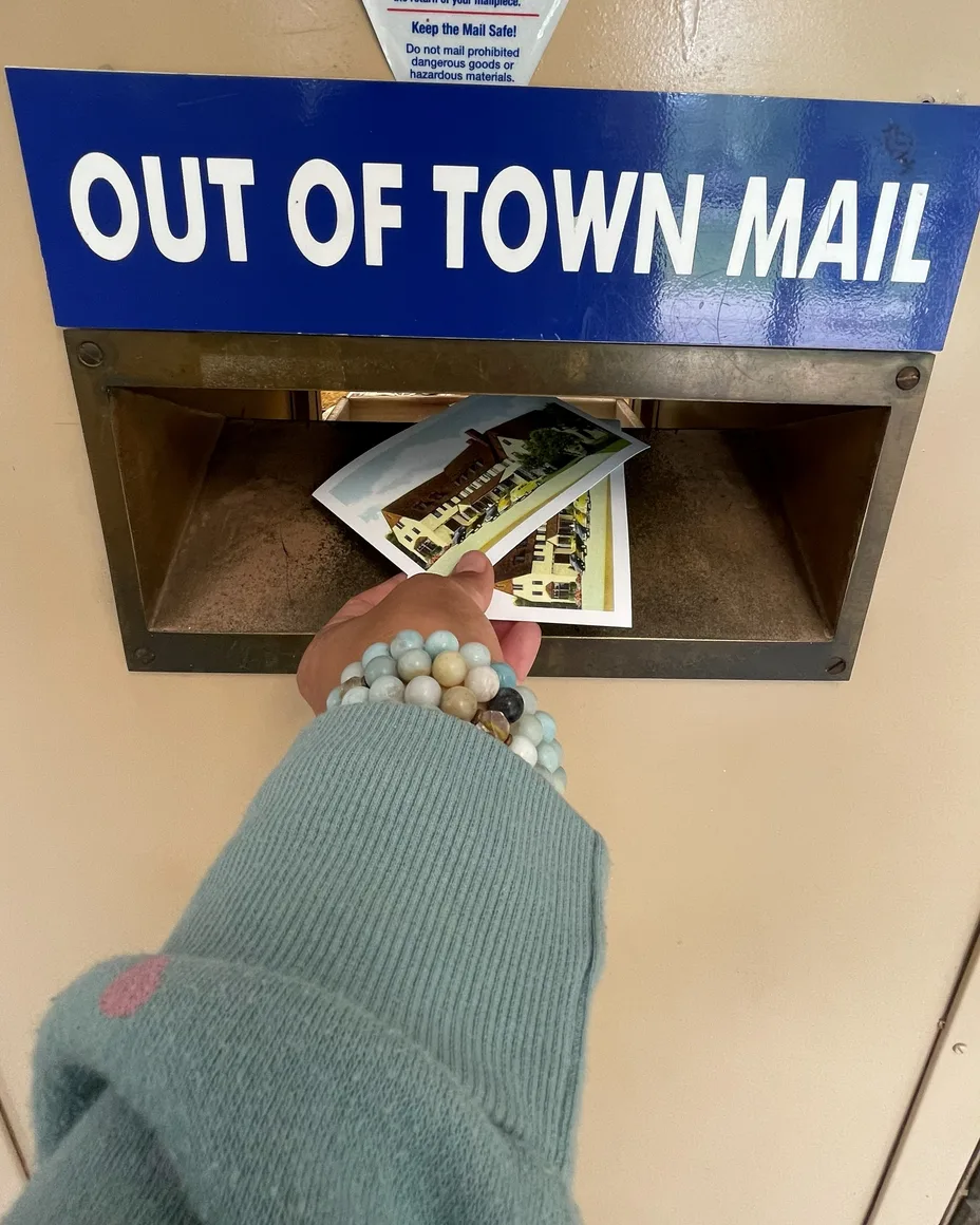 Her hand, in a green sweater with red hearts, dropping postcards into the Out of Town Mail slot.