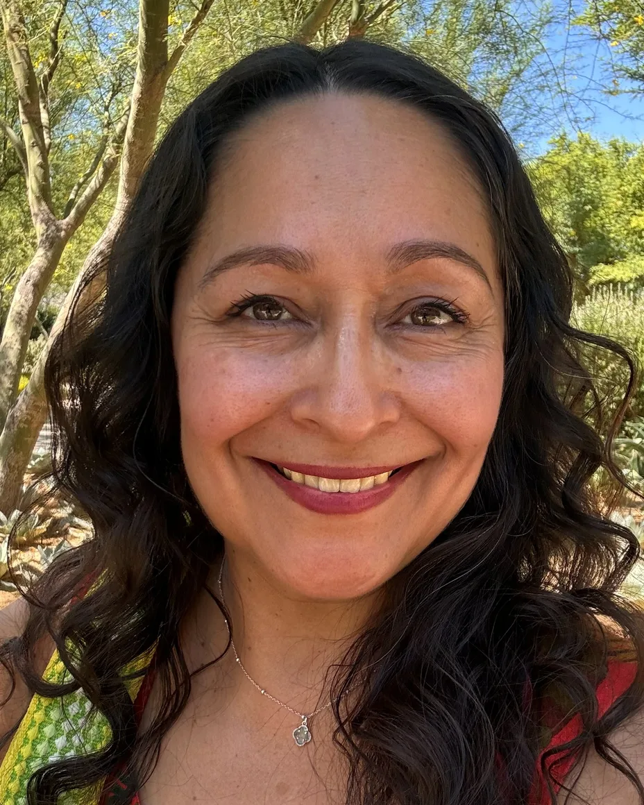 Nikki smiling in the sun, her dark hair loose, in a red dress with green trees behind her.