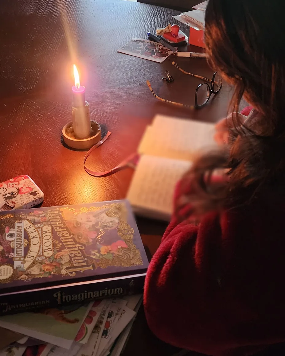 Nikki bent over her notebook in a red robe, a candle and her glasses on the table.
